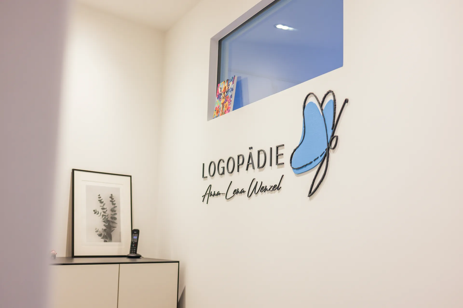 Das Logo der Praxis an der Bürowand: der Schriftzug Logopädie Anna-Lena Wenzel und ein blauer Schmetterling, daneben ein Sideboard