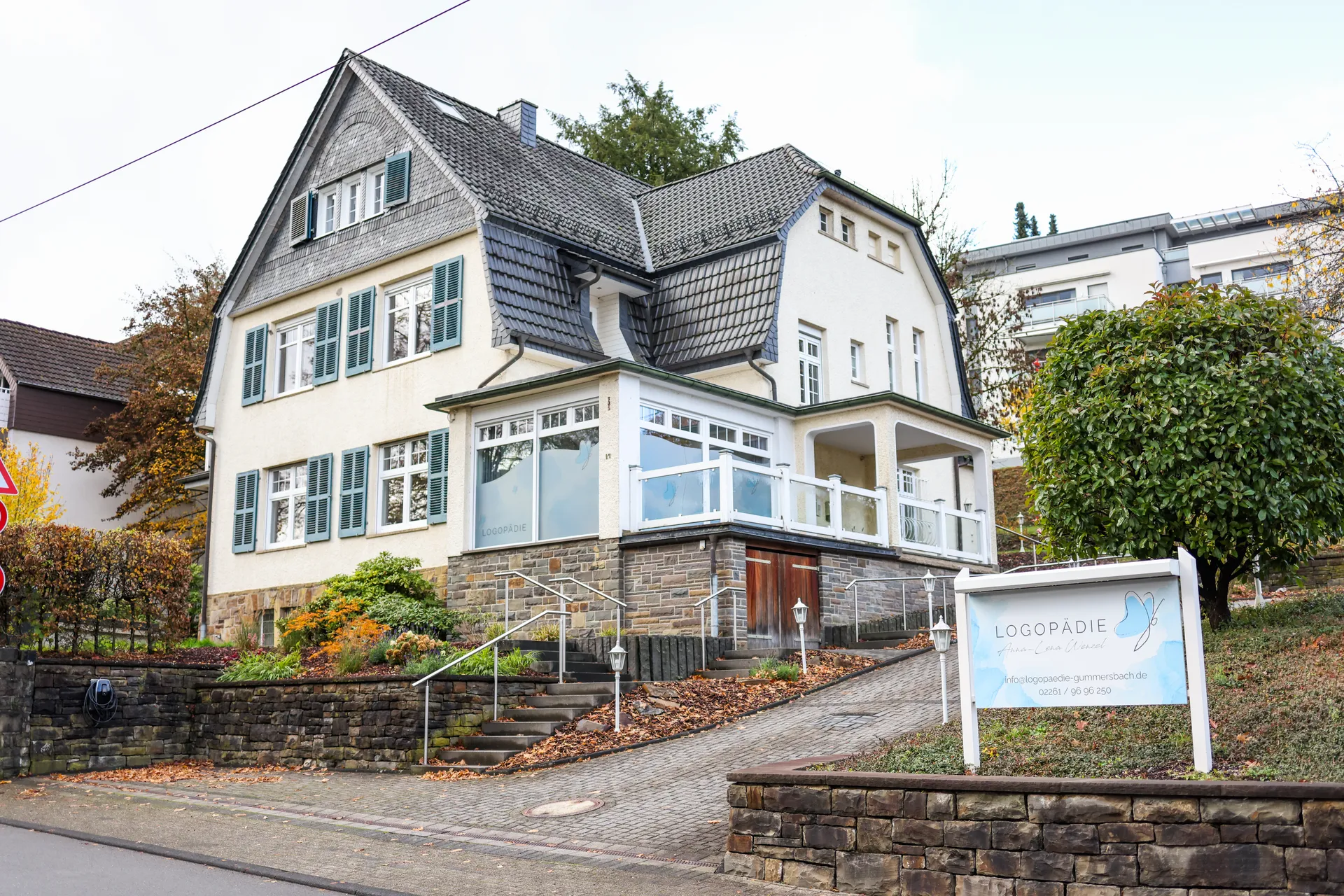 Die Praxis liegt in einer Altbauvilla; von der Straße führen Treppen hinauf, am Fußweg steht das Praxisschild. Rechts neben dem Haus liegen sechs eigene Parkplätze, auf diesem Bild vom Baum verdeckt