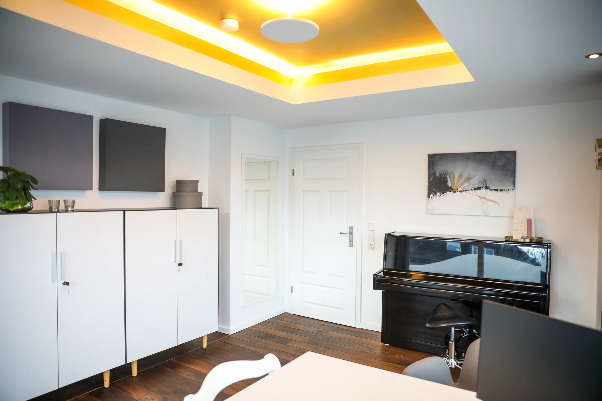 Therapieraum 1 von der anderen Seite: weiße Schränke, ein Klavier und indirektes Licht in der Deckenkehle