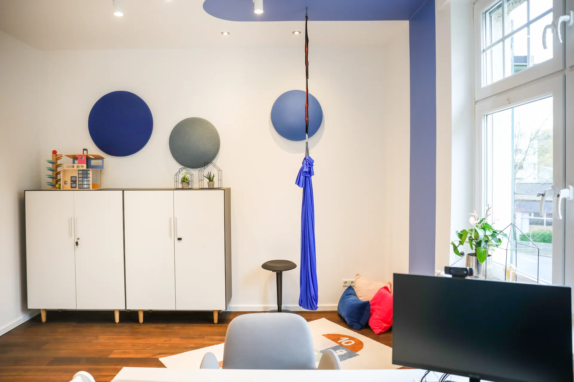 Therapieraum 2 mit runden blauen Akustikpaneelen an der Wand, einem blauen Hängetuch von der Decke und einem grau-weißen Sideboard