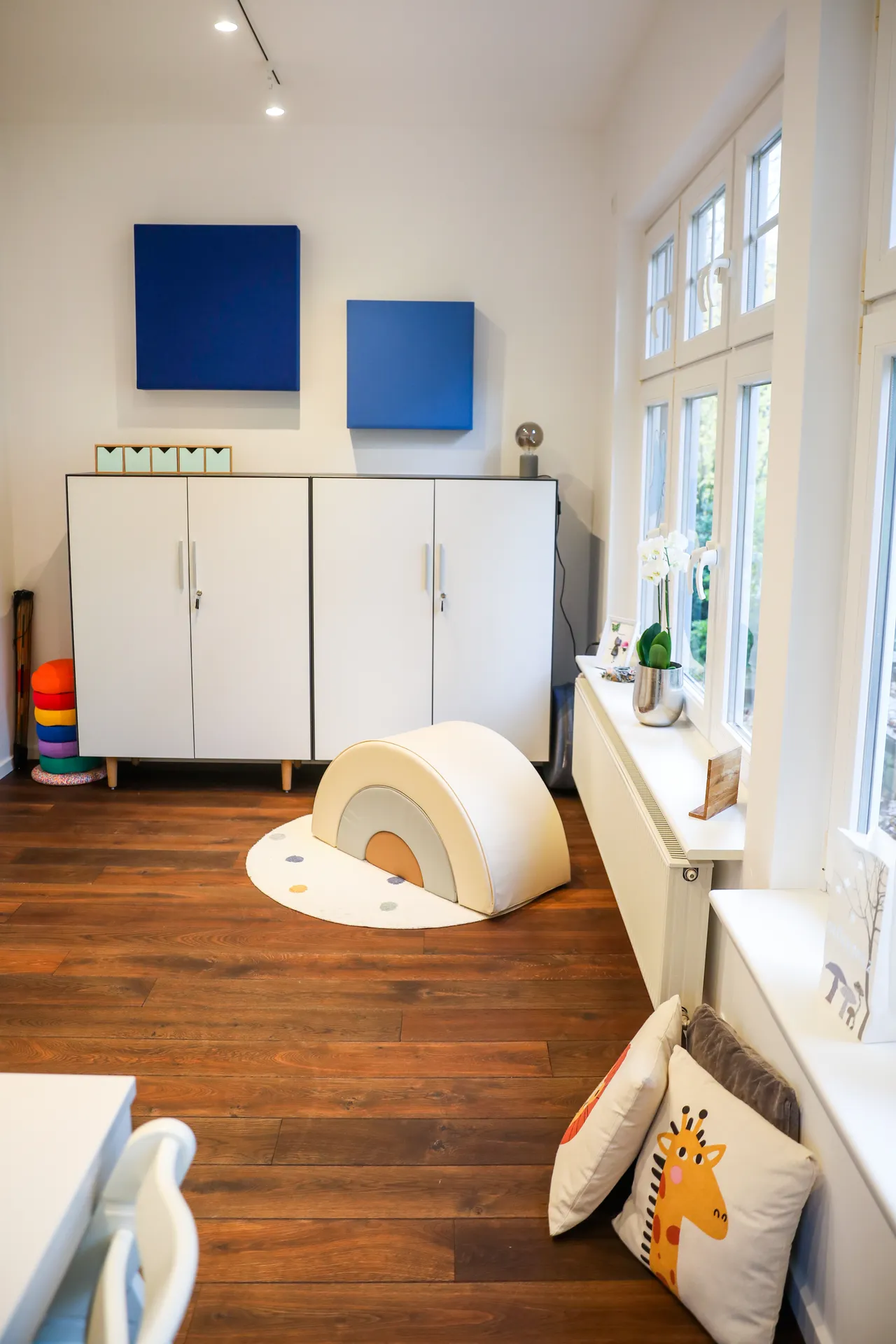 Therapieraum 4 aus der Raummitte: eckige blaue Akustikpaneele an der Wand, weiße Schränke und die Regenbogenwippe
