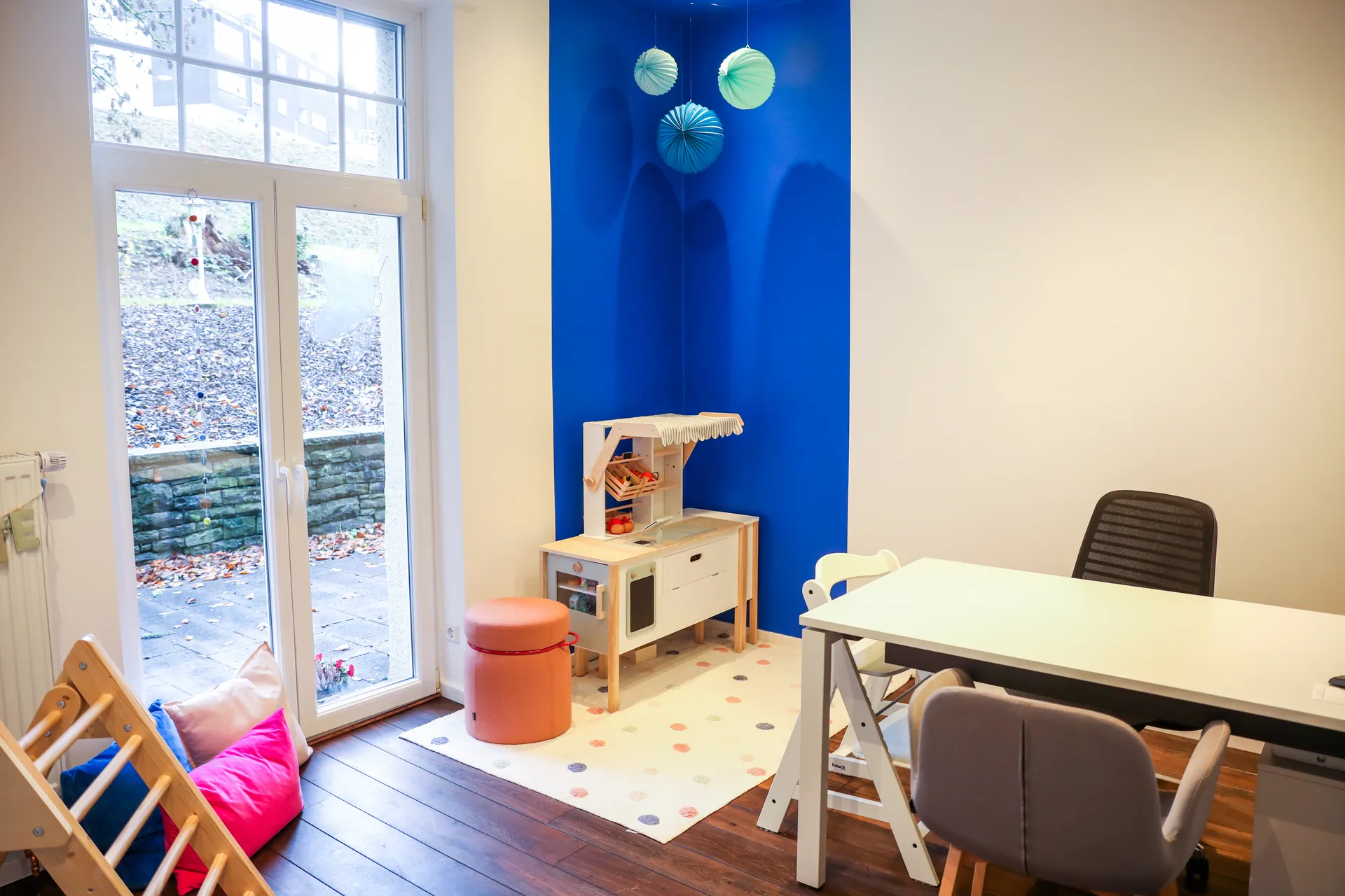 Therapieraum 5 mit Arbeitstisch, Kinderstühlen, einer Spielküche aus Holz und einer Tür zum Garten