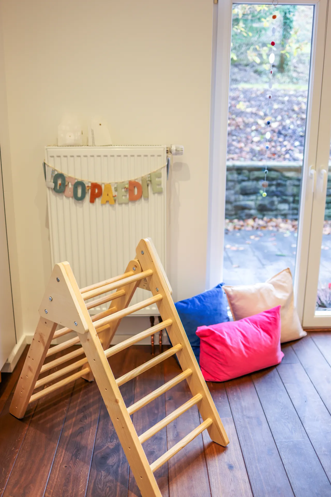 Ein Kletterdreieck aus Holz und Sitzkissen am Fenster, darüber eine Wimpelkette mit dem Wort Logopädie