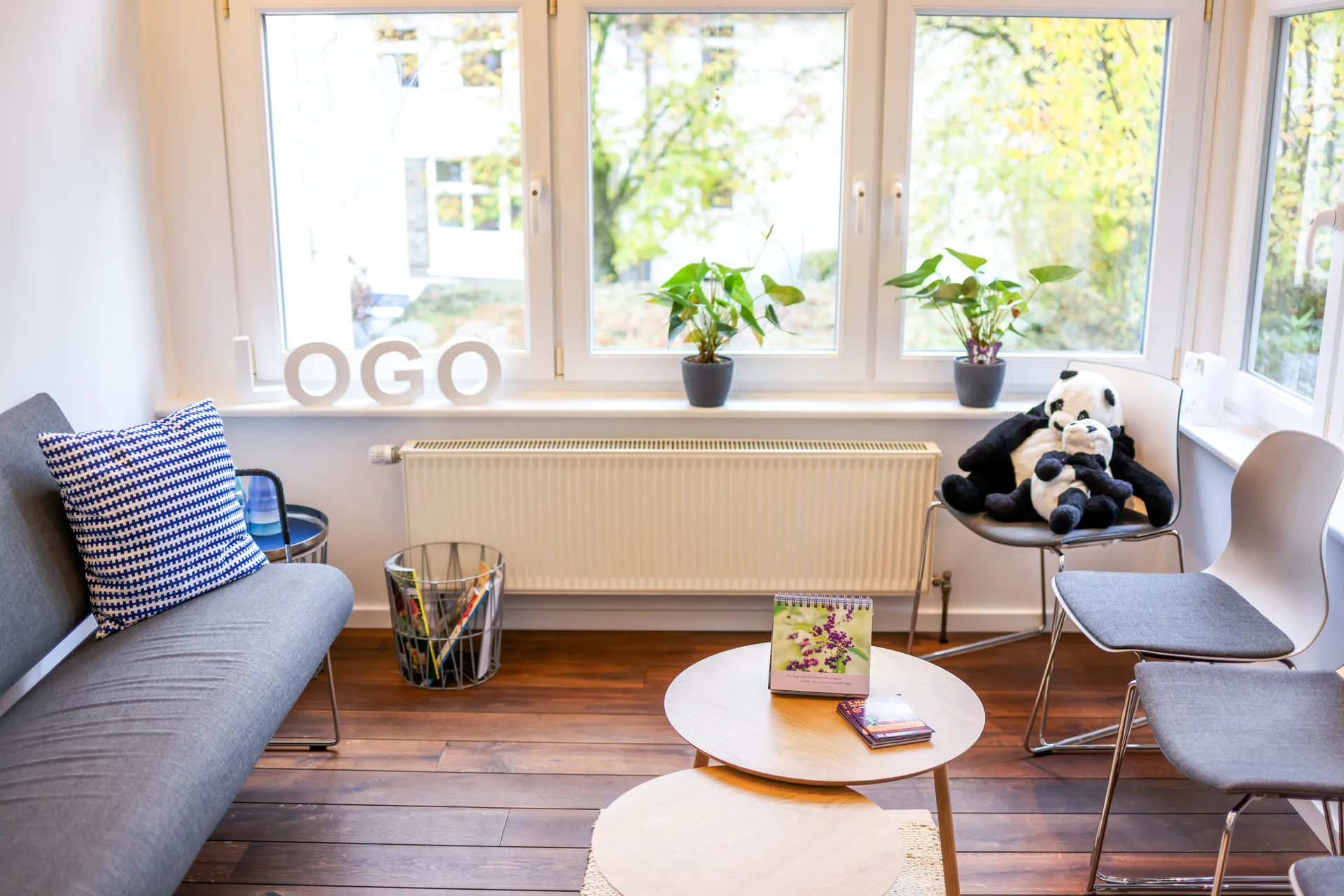 Der Wartebereich mit Sofa, runden Beistelltischen, Stühlen und Pflanzen an der Fensterfront